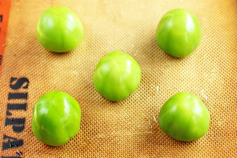 Five green tomatillos with the husks removed arranged on a Silpat nonstick pan liner.