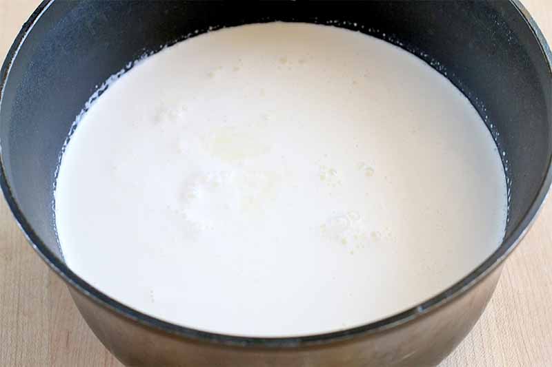 A white milk and cream mixture fills a black sauce pan about halfway, on a beige countertop.