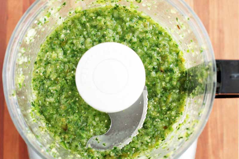 Top-down shot of a food processor with green homemade tomatillo salsa at the bottom, on a wood background.