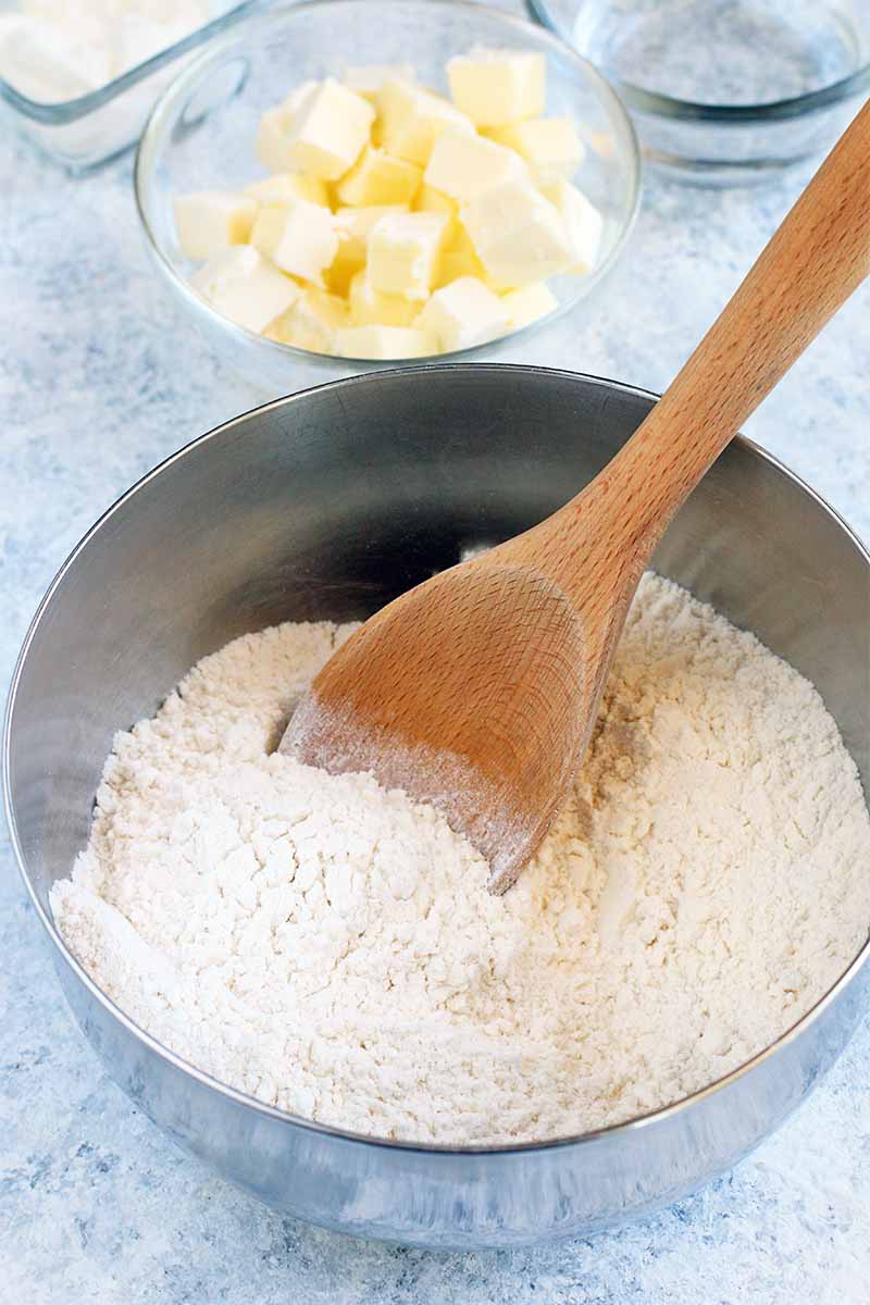 A wooden spoon stirs a flour mixture in a stainless steel mixing bowl, beside small glass bowls of cubed butter and water.