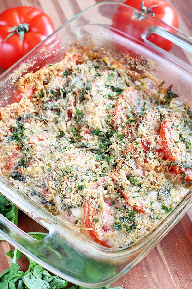 Vertical top-down shot of a square glass baking dish of a casserole with melted cheese, fresh herbs, and golden brown breadcrumbs on top, on a brown wood surface with two red tomatoes and a sprig of green basil.