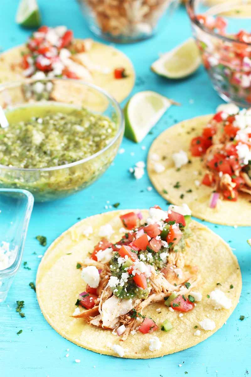 Several corn tortilla topped with shredded chicken, crumbled cheese, and salsa, on a bright blue background with bowls of salsa and spices, and scattered lime wedges and chopped fresh herbs.