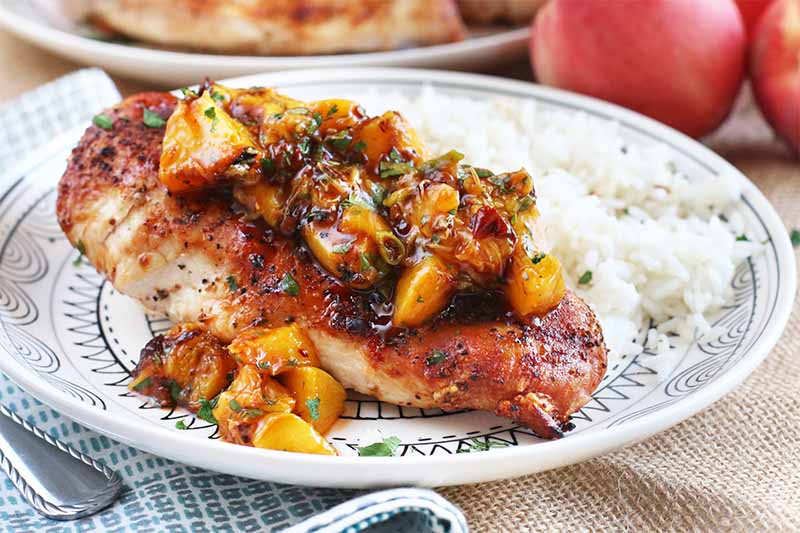 Closeup of a grilled chicken breast topped with peach salsa, beside a small pile of white rice, on a folded cloth napkin with a fork, with more whole fruit in the background.