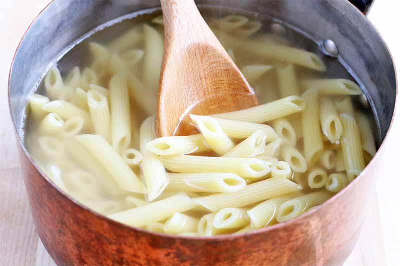 Cooked pasta in water in a copper colored saucepan, with a wooden spoon, on a beige countertop.