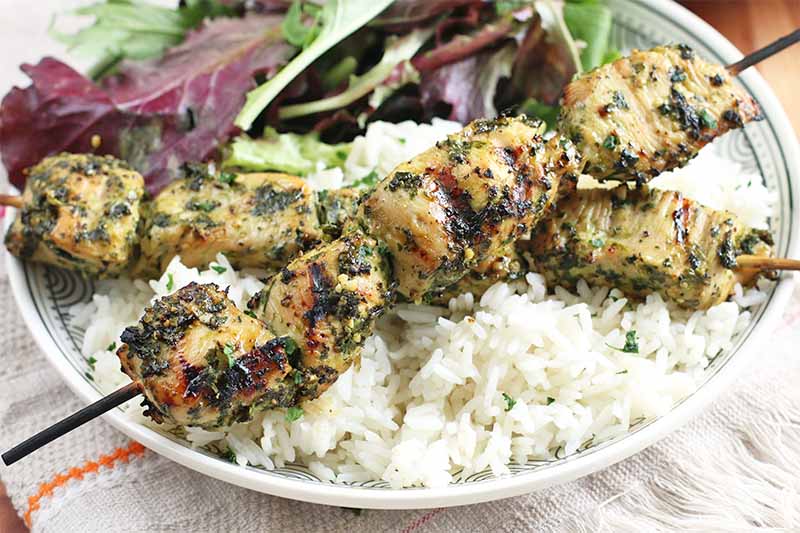Two honey and lime marinated chicken skewers on a bed of white rice, beside a small pile of salad greens on a white plate.