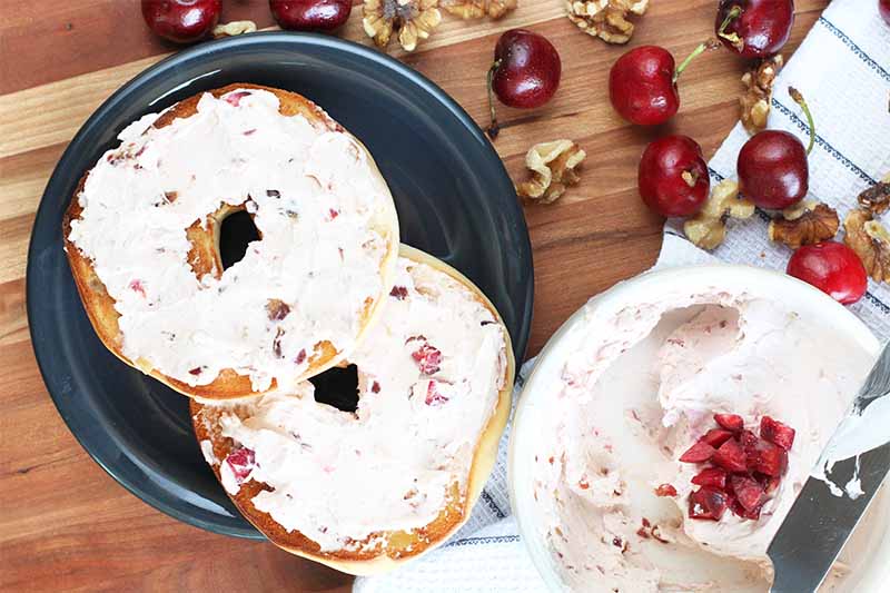 Horizontal image of bagel slices spread with cream cheese surrounded by nuts and whole cherries.