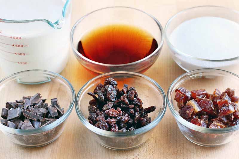 A glass pitcher measuring cup of milk and five small glass bowls of spiced rum, heavy cream, chocolate chunks, raisins, and chopped dates, arranged in two rows on a beige countertop.