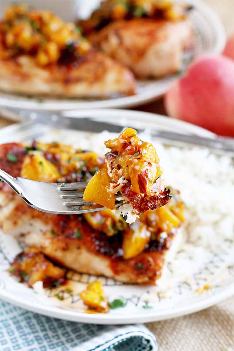 A forkful of chicken with peach chipotle sauce in the foreground, with more on two plates with white rice in the background, and a few whole stone fruit in soft focus.