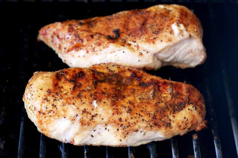Two whole chicken breasts rubbed with spices, cooking on a grill.