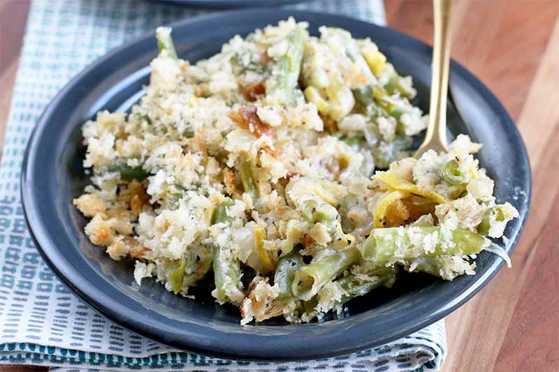 A blue plate filled with a creamed green bean and yellow squash casserole, topped with roasted breadcrumbs, with a fork, on top of a folded blue and white checked cloth napkin, on a brown wood table.