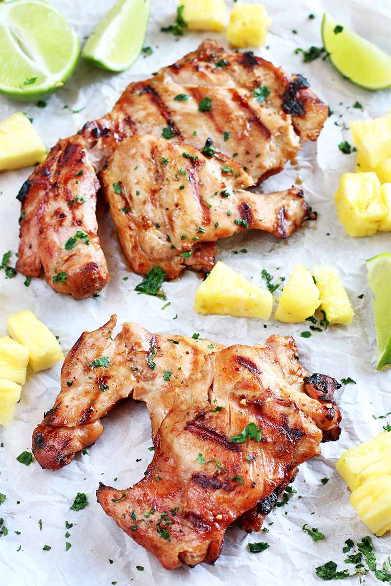 Two boneless skinless grilled chicken thighs on a piece of white parchment paper with yellow pineapple chunks, lime wedges, and a spinkling of fresh cilantro.