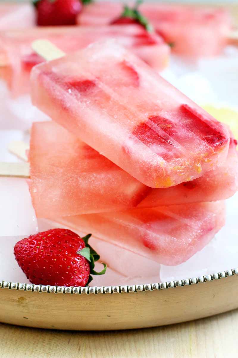 A stack of three strawberry lemonade popsicles on a sterling silver tray, with a whole berry and more fruit and freeze pops in the background.