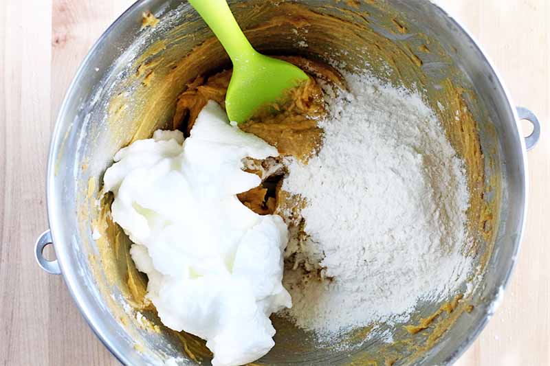 Top-down shot of a thick beige batter, whipped egg whites, and a flour mixture in a stainless steel mixing bowl with a lime green silicone spatula.