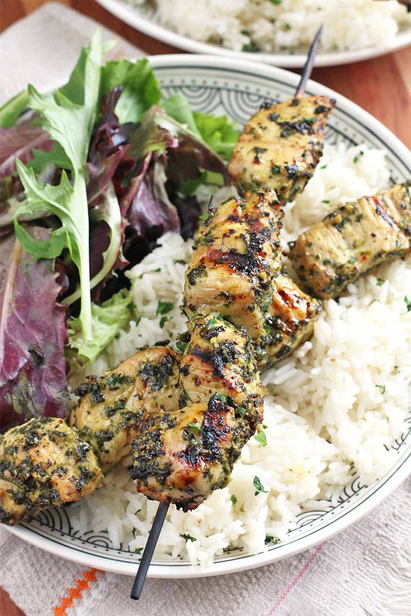 Two herb marinated and grilled wooden skewers of chicken breast chunks, on a bed of white rice next to a small pile of salad greens, on a white plate, on top of a folded white cloth with an orange stripe, with another identical dish in the background.