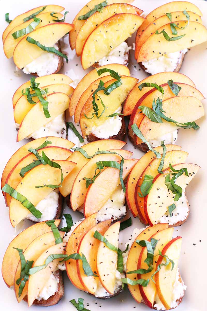 Top-down shot of a grid of three by four small appetizer toasts topped with ricotta, peach slices, and fresh basil chiffonade.