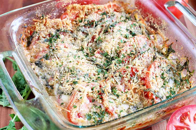 A baked tomato dish topped with chopped green herbs and golden brown melted cheese.