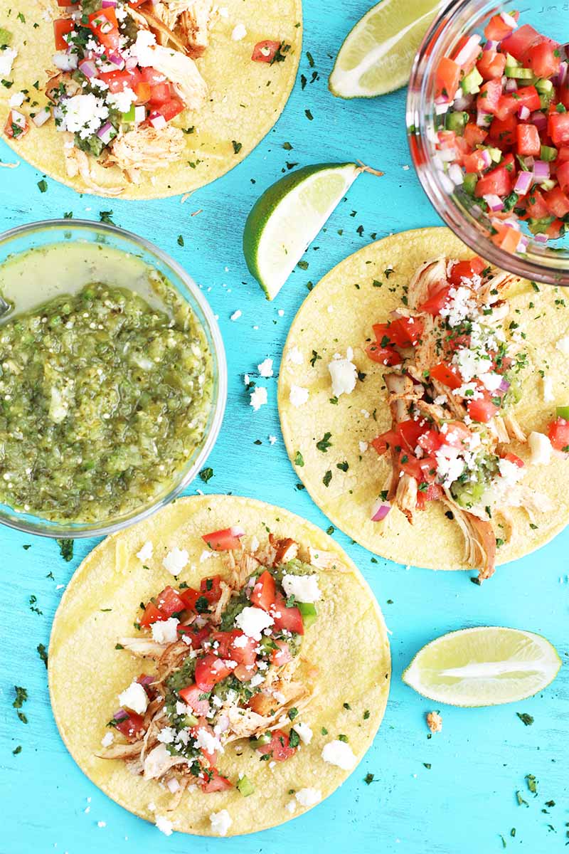 Top-down shot of three round corn tortilla topped with shredded chicken, salsa verde, pico de gallo, and chopped cilantro, next to bowls of more red and green salsa, lime wedges, and scattered chopped herbs on a blue background.