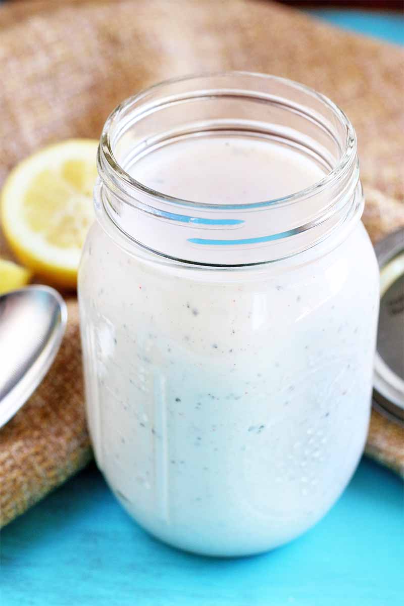 A large mason jar of a thick white liquid, on a bright blue surface with a spoon, metal lid, and cut lemon on top of a piece of burlap in the background.