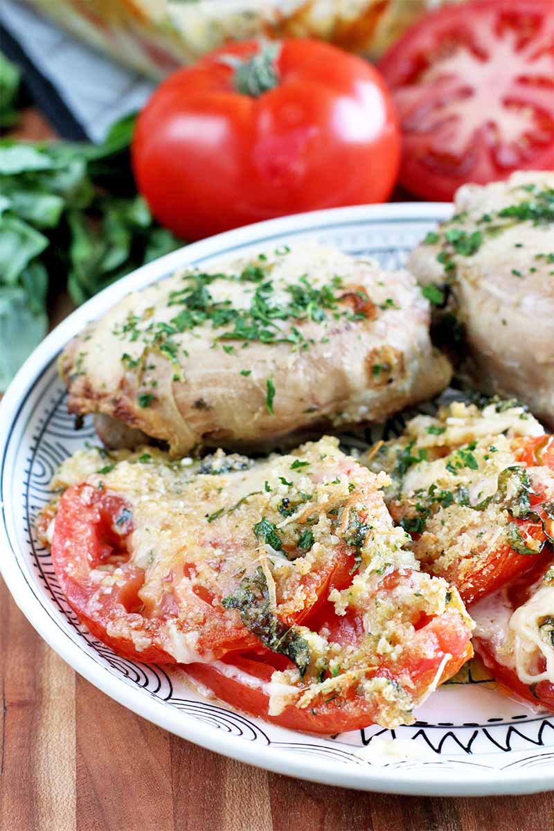 Scalloped tomatoes with chicken parmesan topped with minced parsley on a white and blue plate, with whole, fresh vegetables and herbs in the background.