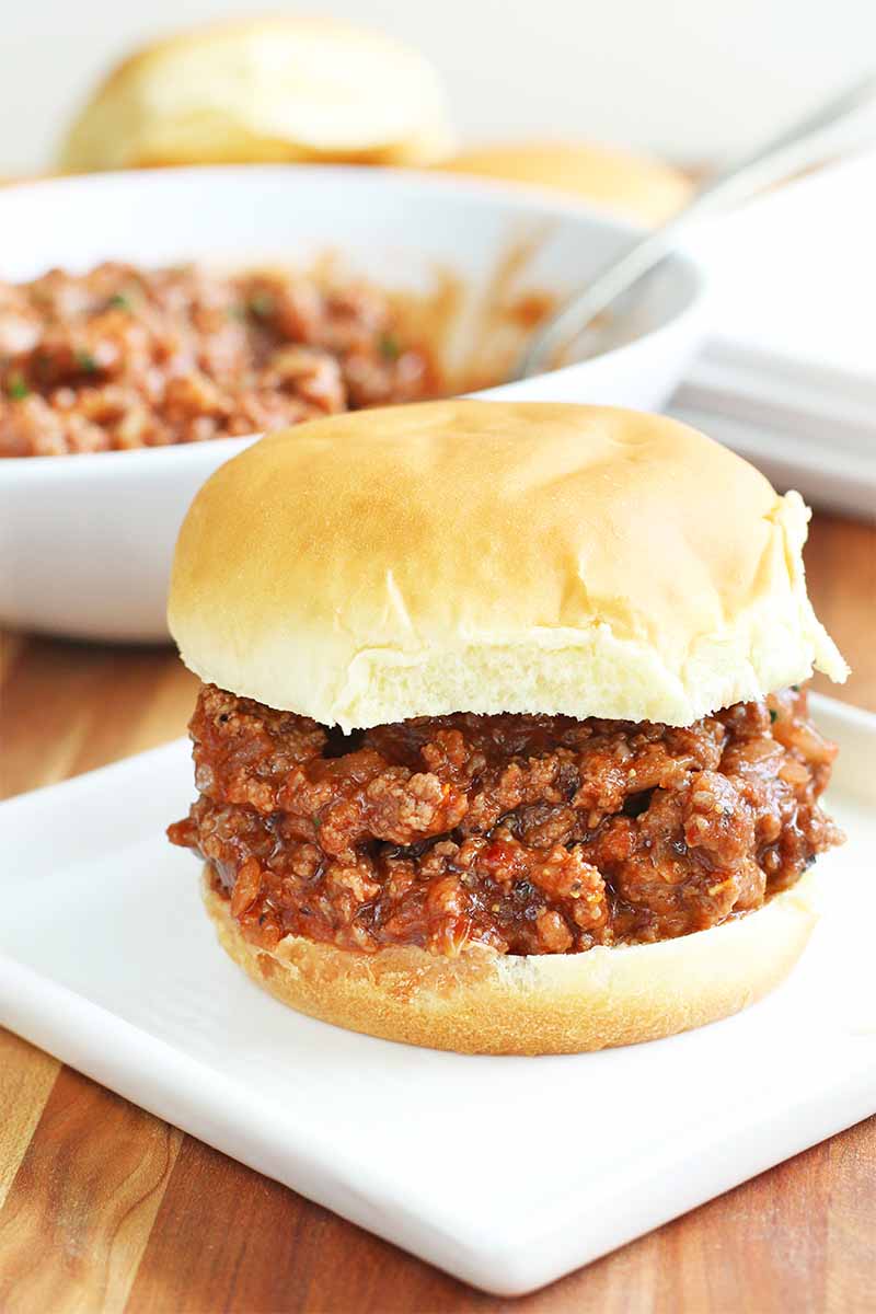 Homemade sloppy joe mixture on a hamburger bun, on a square white plate with more in a white bowl with a spoon in the background, on a brown wood surface.