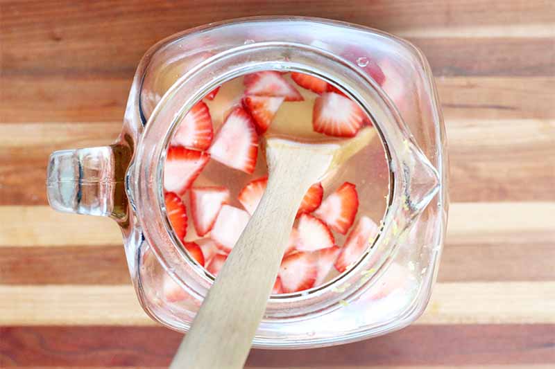 Top-down shot of a clear glass pitcher of strawberries and lemonade, with a wooden spoon, on a striped wooden surface.