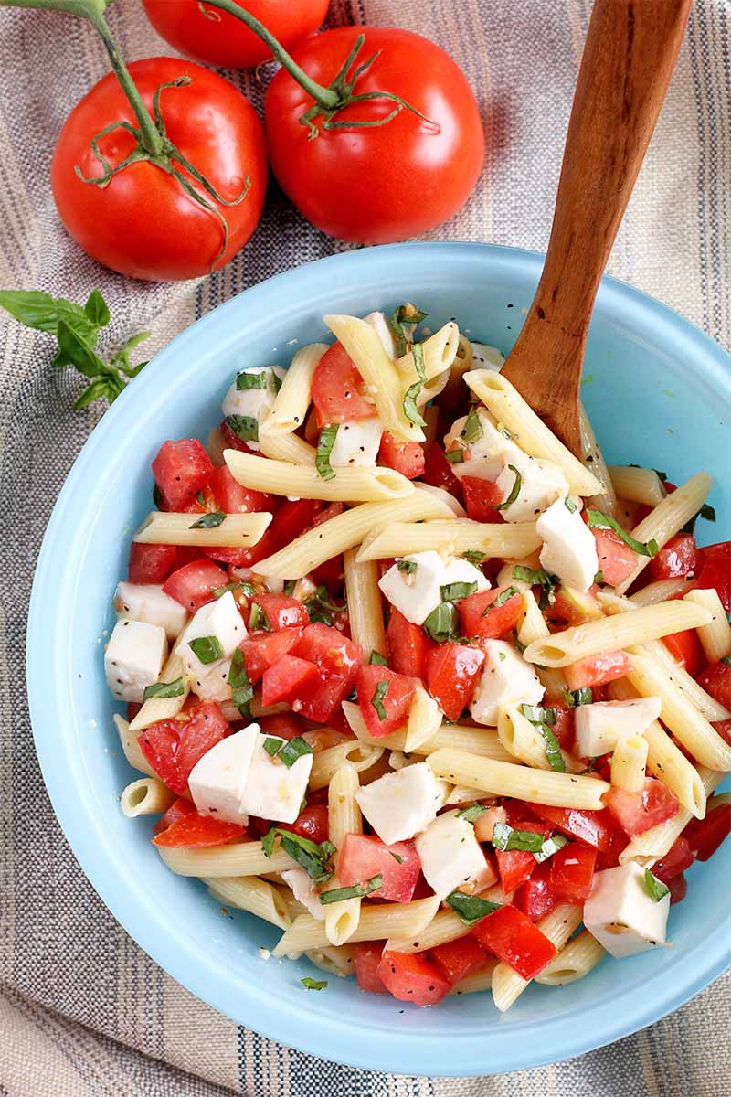 Top-down shot of a light blue glass bowl of pasta salad with a wooden spoon, on a folded gray cloth with a small sprig of basil and three red tomatoes.
