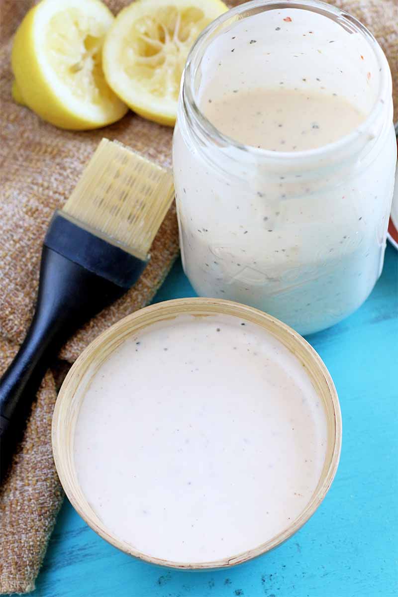 Vertical image of a large mason jar and a small off-white ceramic bowl of white barbecue sauce, beside a black-handled OXO silicone brush with a halved and juiced lemon on a piece of burlap, on a bright blue background.