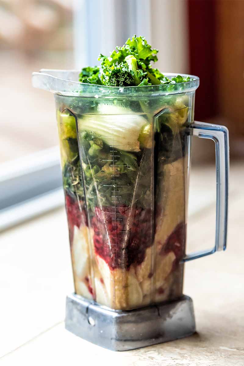 An overfilled plastic blender canister, with kale and a variety of vegetables, fruits, and berries, on a white countertop in front of a window.