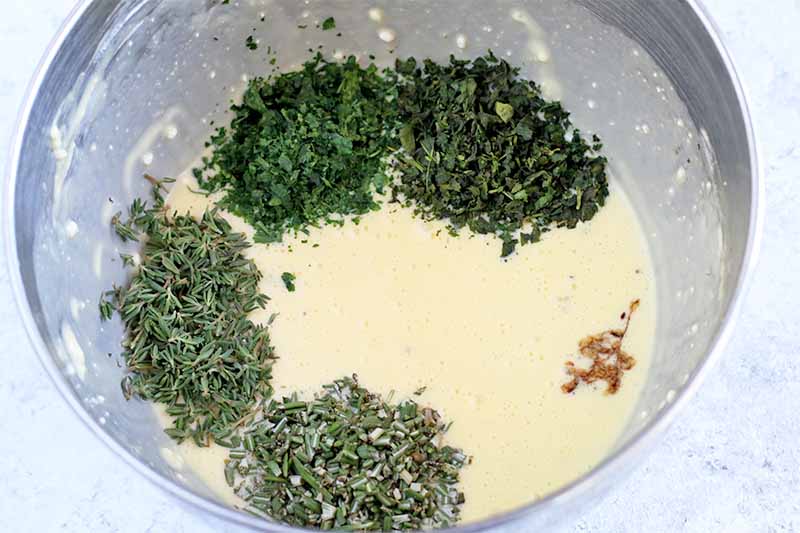 Four small piles of different types of green herbs and a drizzle of balsamic vinegar on top of an egg mixture in a stainless steel mixing bowl.