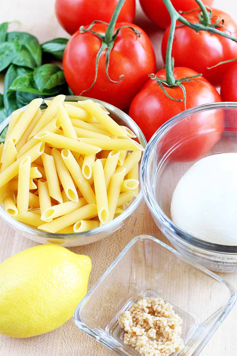 Bowls of uncooked penne, fresh mozzarella, and minced garlic, with a yellow lemon, a bunch of green basil, and a cluster of six red tomatoes on the vine, on a beige countertop.