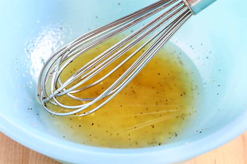 A wire whisk rests in a light blue glass bowl, with a yellow vinaigrette mixture at the bottom, on a beige countertop.