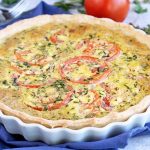 Tomato, egg, and herb tart in a white ceramic pan, on a blue cloth with scattered ingredients.