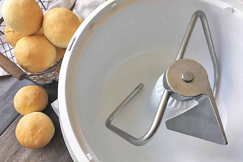 Horizontal image of a dough hook in a plastic bowl next to baked bread rolls.