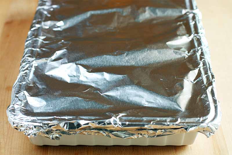 Closely cropped oblique image of a foil-wrapped baking dish, on a beige background.