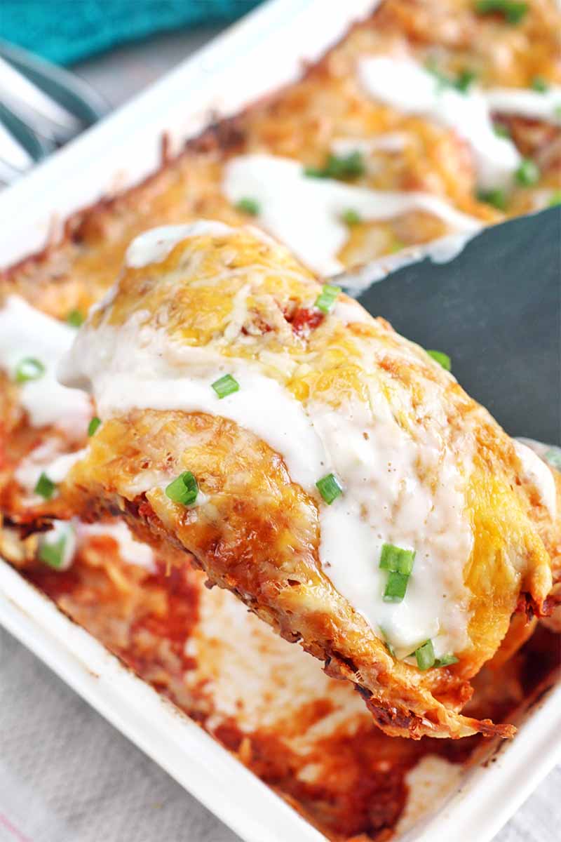 A metal spatula holds a chicken enchilada topped with blue cheese dressing and chopped scallions up to the camera, with the rest of the casserole in a white ceramic baking dish in the background.