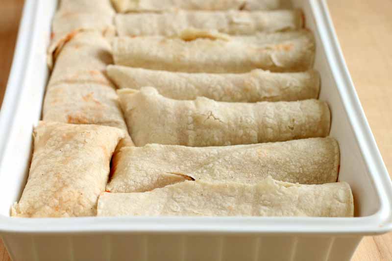Rolled tortillas fill a white, rectangular, ceramic baking dish, on a beige background.