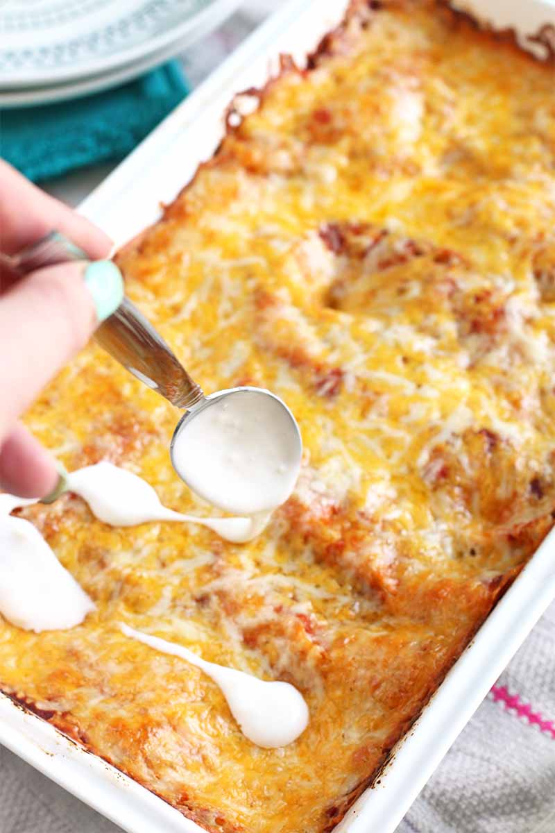 A woman's hand with light blue manicured nails drizzles blue cheese dressing onto the top of a casserole with melted cheese covering the surface.
