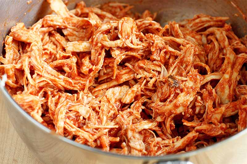 Shredded chicken coated with red sauce in the bottom of a stainless steel bowl, on a white background.