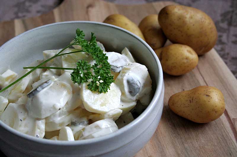 Close up of white porcelain bowl full of German potato salad garnished with parsley with raw potatoes in the background. 