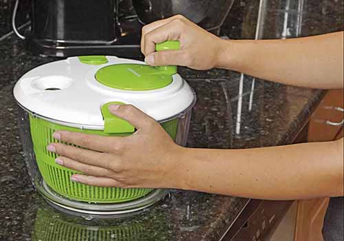A person holds a green and white plastic Cuisinart salad spinner still on a black granite kitchen countertop with one hand, and turns the crank on top with the other.