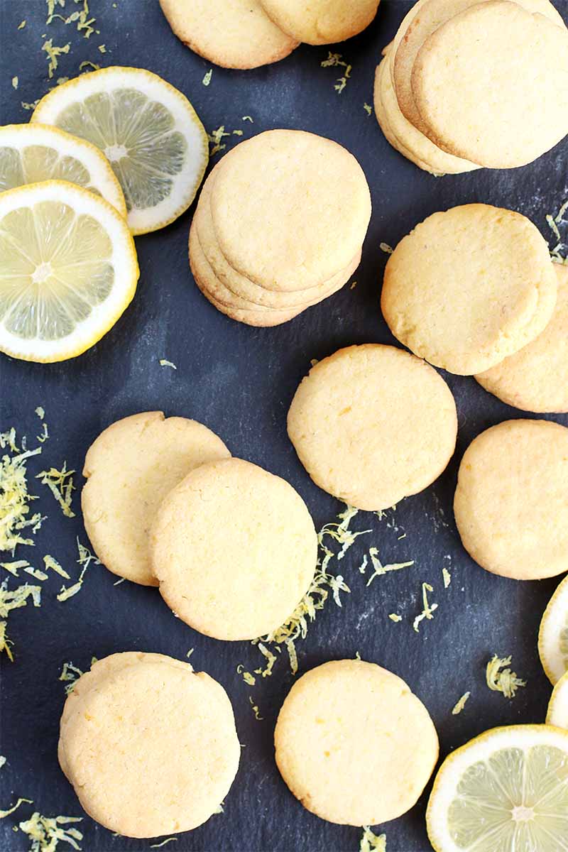 Top-down shot of informally arranged butter cookies with scattered lemon slices and zest on a black slate background.