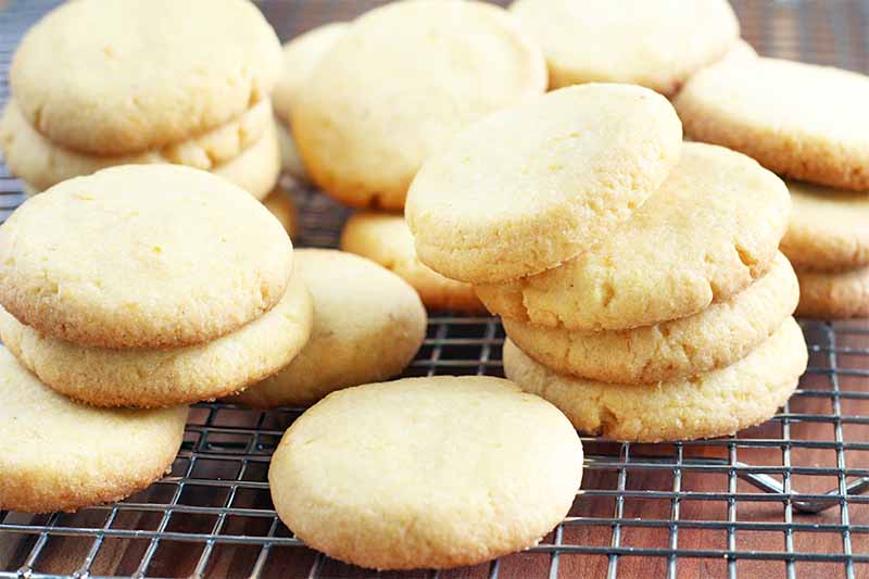 Lemon cornmeal cookies are arranged haphazardly in short stacks on a wire rack, on a brown background.