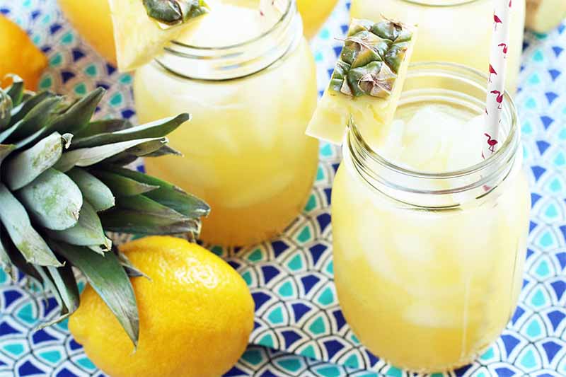 Three glass mason jars of tropical lemonade, with pink and white paper straws, a pineapple top and pieces arranged on the lip of each jar for garnish, and a whole lemon, on a light and dark blue patterned background.