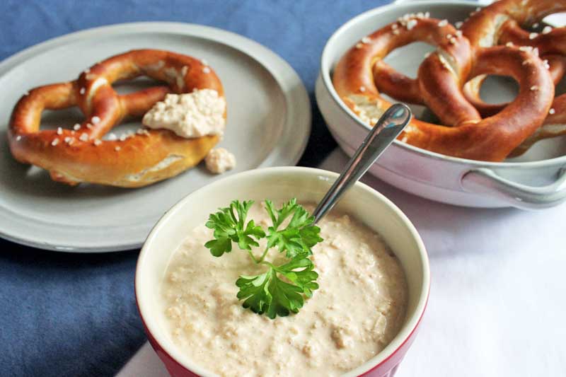 A bowl of German Bavarian Obatzda cheese dip in the center front and two plates with large lye pretzels in the rear. On a blue tablecloth.