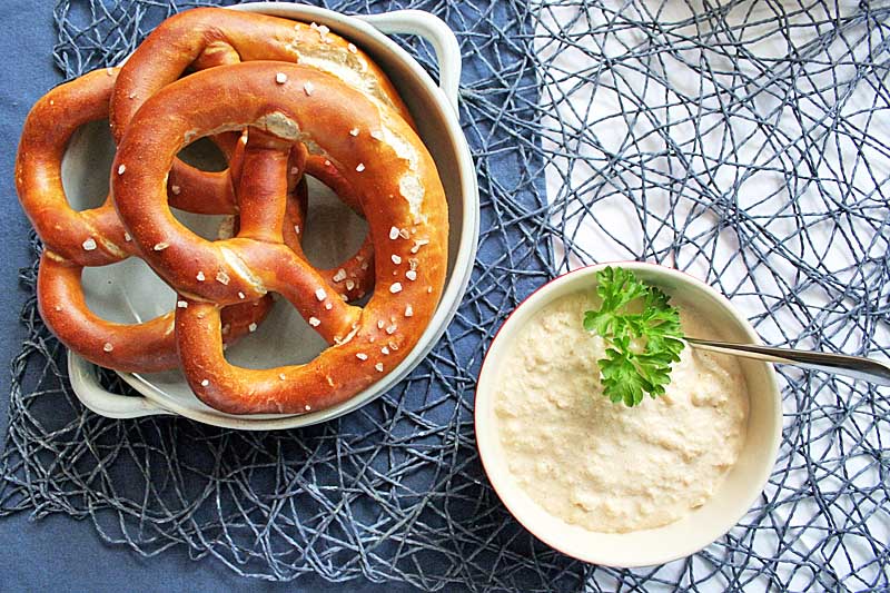 Top down view of a bowl containing large lye pretzels on the upper left with a bowl of German Obatzda cheese dip on the lower right.
