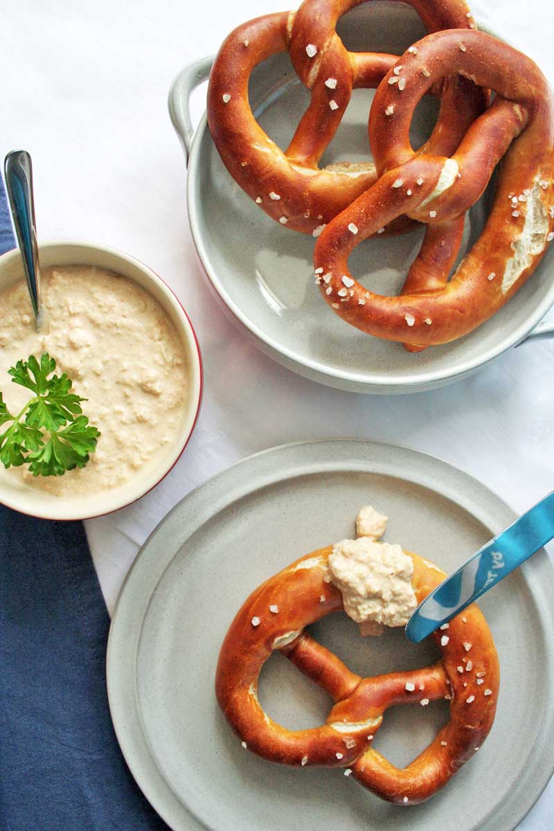Top down view of a three lye pretzels on white, porcelain plates with a a bowl of German Obatzda cheese dip placed to the left.