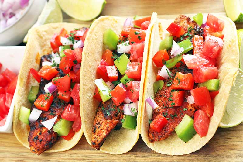 Three Cajun chicken tacos topped with chopped vegetables, on a wood cutting board with small ceramic bowls of garnish and lime wedges.