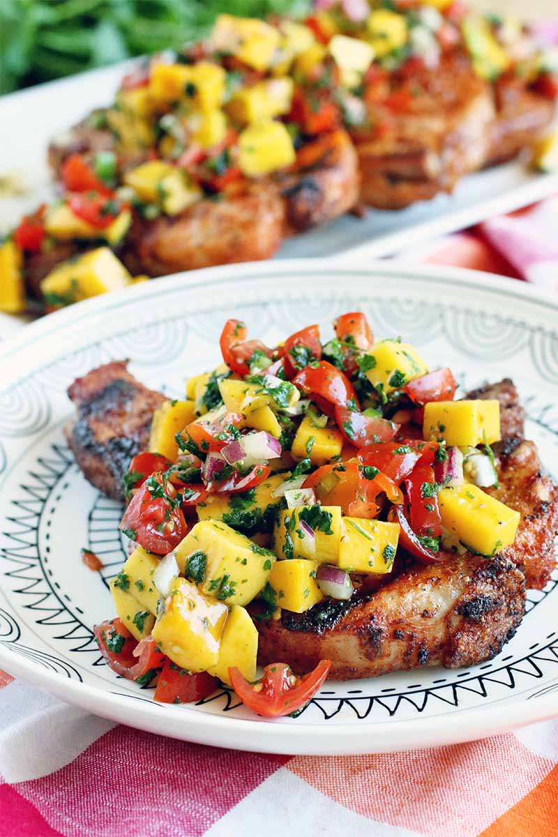 A grilled pork chop topped with fruit salsa on a black and white plate, with more on a rectangular serving plate in the background, on a red, orange, and white checkered tablecloth with green cilantro in shallow focus in the background.