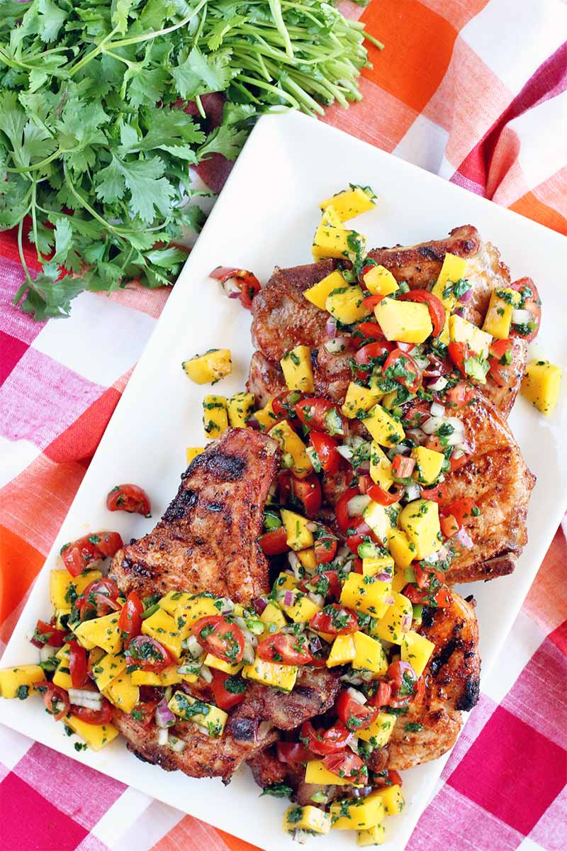 Top-down shot of four spice-rubbed and grilled bone-in pork chops on a rectangular white serving plate, topped with fresh mango salsa, next to a bunch of green cilantro on a red and white checkered tablecloth.