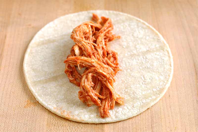 A pile of shredded chicken coated in a red sauce is arranged in a line at the center of a corn tortilla, on a beige particleboard background.